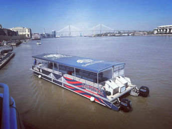 Blue-canopied riverboat on the Savannah River with a cable-stayed bridge and waterfront skyline under a clear blue sky