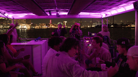 Nighttime river cruise party under purple LED canopy with passengers chatting and singing karaoke, city waterfront and illuminated bridge in the background.
