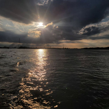 Sun breaking through dramatic storm clouds, golden rays reflecting on rippled water leading to a silhouetted industrial shoreline with cranes and a distant bridge at sunset.