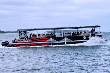 Open-sided sightseeing pontoon boat full of passengers speeding across coastal waters, leaving a choppy wake with low marshy shoreline and overcast sky in the background.