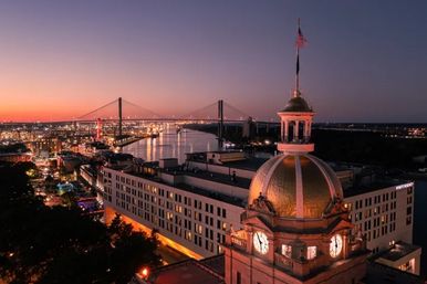 Savannah, Georgia skyline at dusk with a gold-domed historic clock tower in the foreground and the Talmadge Memorial Bridge spanning the river under a colorful sunset