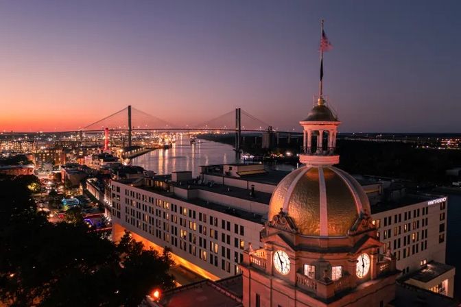 Savannah, Georgia skyline at dusk with a gold-domed historic clock tower in the foreground and the Talmadge Memorial Bridge spanning the river under a colorful sunset