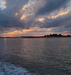 Vibrant sunset over coastal marshland, dramatic clouds and orange-pink light reflecting on rippling water with a boat wake in the foreground.
