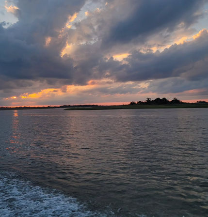 Vibrant sunset over coastal marshland, dramatic clouds and orange-pink light reflecting on rippling water with a boat wake in the foreground.