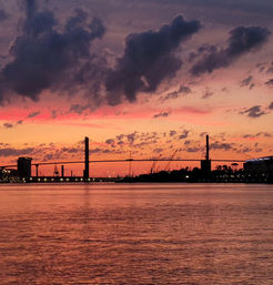 Vibrant pink-orange sunset over a cable-stayed bridge and harbor cranes, calm river reflecting colorful sky