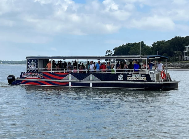 Open-sided river tour boat carrying a crowd of passengers cruising a calm coastal river past tree-lined shoreline and waterfront homes under a partly cloudy sky.