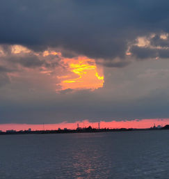 Glowing orange sunset punching through dark storm clouds over a calm bay with a silhouetted shoreline and distant bridge