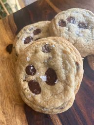 Close-up of freshly baked chocolate chip cookies with melted chocolate chips and flaky sea salt on a wooden cutting board