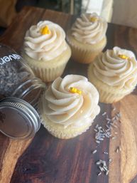 Four vanilla cupcakes with swirled ivory buttercream and tiny yellow sugar accents arranged on a rustic wooden board with scattered lavender buds and a tipped glass jar nearby
