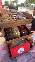 Assorted oversized cookies — chocolate chip, red velvet, double chocolate and filled varieties — arranged on a wooden tiered display at an outdoor market stall.