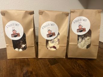 Three kraft paper bakery bags with round white labels and clear windows showing chocolate and white chocolate bark pieces with mix-ins, arranged on a wooden table