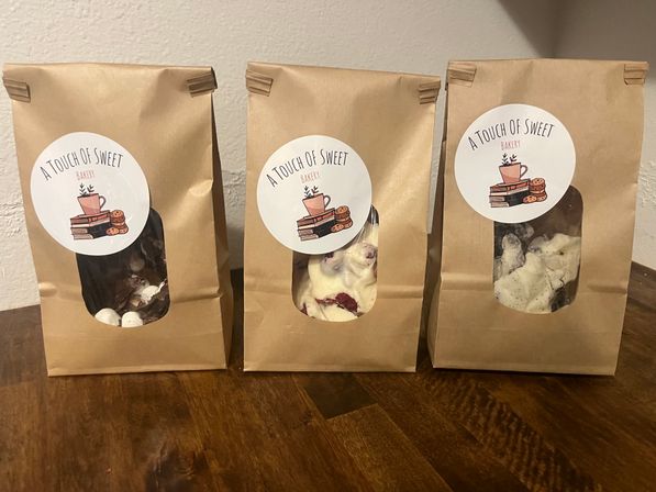 Three kraft paper bakery bags with round white labels and clear windows showing chocolate and white chocolate bark pieces with mix-ins, arranged on a wooden table