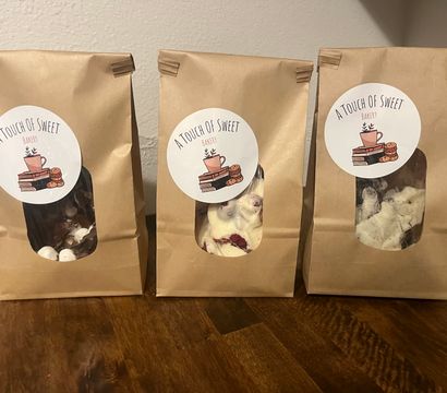 Three kraft paper bakery bags with round white labels and clear windows showing chocolate and white chocolate bark pieces with mix-ins, arranged on a wooden table