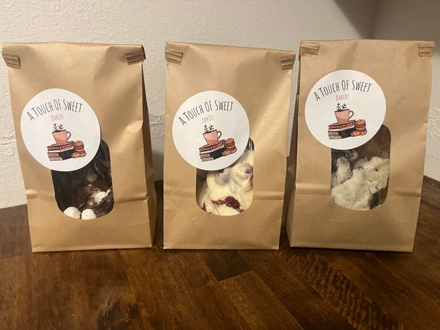 Three kraft paper bakery bags with round white labels and clear windows showing chocolate and white chocolate bark pieces with mix-ins, arranged on a wooden table