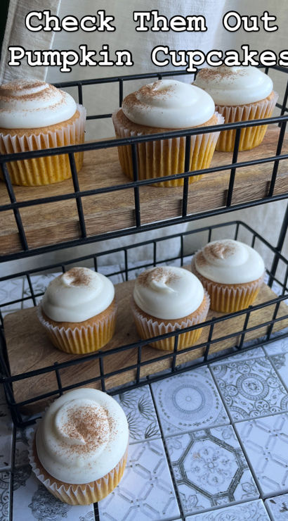 Rustic two-tier wire stand of pumpkin cupcakes topped with creamy swirls of cream-cheese frosting and a cinnamon dusting on patterned tile countertop — bakery-style display.