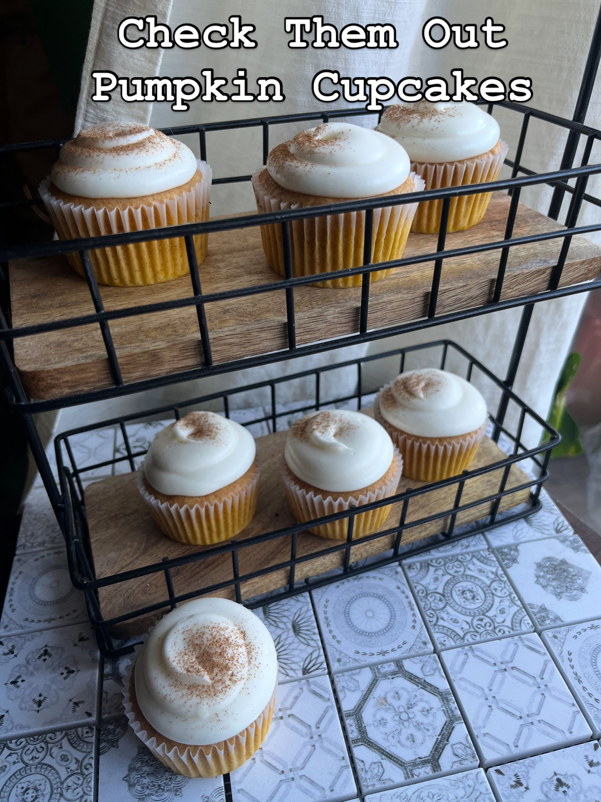 Rustic two-tier wire stand of pumpkin cupcakes topped with creamy swirls of cream-cheese frosting and a cinnamon dusting on patterned tile countertop — bakery-style display.