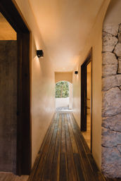Cozy sunlit interior corridor in a modern-rustic tropical home — dark wooden plank floor, smooth beige plaster walls, black cube wall sconces and a textured stone accent wall, leading to an arched opening with a leafy garden view.