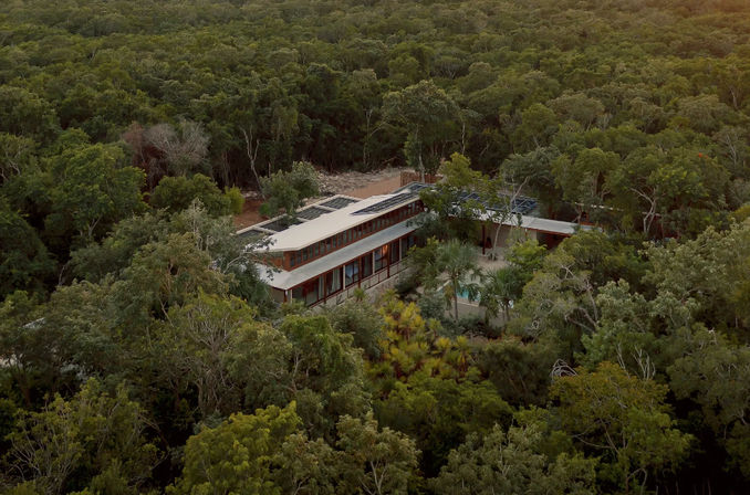 Aerial view of a long modern eco-friendly house with rooftop solar panels and a small pool, nestled in a dense green forest canopy