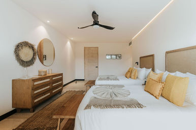 Bright coastal-inspired bedroom with two beds, mustard-yellow pillows, woven headboards, wooden dresser and round mirror, black ceiling fan, and natural-fiber rug.