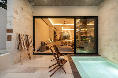Modern villa terrace with private illuminated plunge pool, wooden folding chairs on stone tile flooring, striped towels on a bamboo ladder, and wide sliding glass doors opening to a warmly lit open-plan living room.