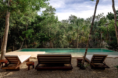 Three wooden sun loungers on a pebble patio facing a turquoise infinity pool set against a lush tropical forest of palms and trees, an inviting secluded jungle oasis.