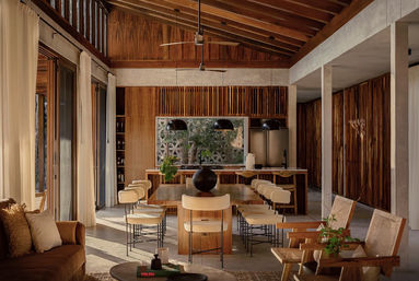 Sunlit open-plan modern-rustic dining and kitchen with vaulted wood ceiling, long wooden table surrounded by cushioned stools, black pendant lights, floor-to-ceiling glass doors and indoor greenery.