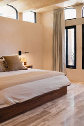 Sunlit minimalist bedroom with low wood platform bed, linen bedding and tassel pillows, warm beige plaster walls, black-framed arched windows, floor-length curtains and polished stone floor.