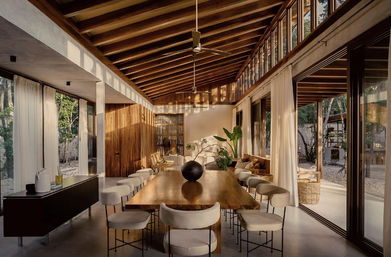 Sunlit modern tropical villa dining room with high exposed wood-beam ceiling, long live-edge wooden table surrounded by upholstered stools, floor-to-ceiling glass walls and sliding doors opening to a leafy courtyard, neutral palette and large potted plants.
