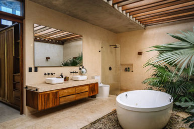 Open-air modern luxury bathroom with round freestanding soaking tub on gravel, dual vessel sinks on a wooden floating vanity, glass shower, large mirror, exposed wood-beam skylight, stone floor and lush tropical palms.