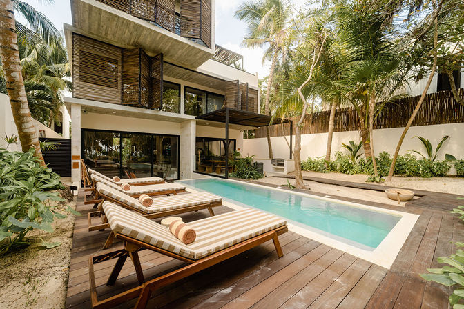 Tropical modern villa pool deck with sun-ready striped lounge chairs beside a rectangular turquoise plunge pool, framed by palm trees, wooden decking, and lush greenery.