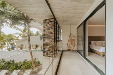 Tropical beachfront balcony with glass railing and hanging rattan swing chair, sliding glass doors to a modern bedroom, palm trees and pool visible beyond.