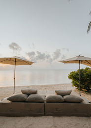 Two oversized cushioned beach loungers beneath matching umbrellas on a sandy tropical beach, calm ocean and pastel sunset sky with palm fronds and greenery at the edges.