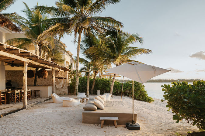 Tropical beachfront lounge with palm trees, white sand, cushioned daybed and umbrella beside the calm ocean at golden hour