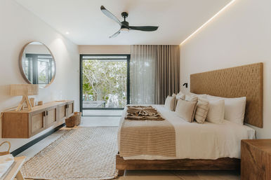 Airy modern bedroom with king bed, woven headboard and layered beige linens, wood floating console with round mirror, ceiling fan, jute rug and sliding glass doors opening to a leafy patio — neutral, coastal-inspired design.