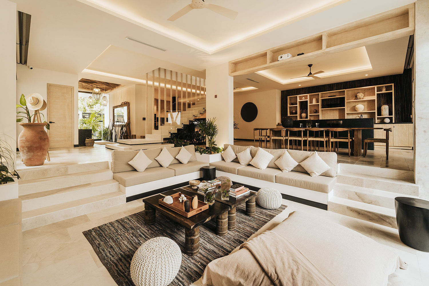 Sunlit resort-style open-plan living room with sunken built-in neutral sofa and throw pillows, wooden coffee table on textured rug, round knit poufs, terracotta vase and plants, steps up to a long wooden dining table and open shelving—modern tropical villa interior.