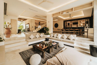 Sunlit resort-style open-plan living room with sunken built-in neutral sofa and throw pillows, wooden coffee table on textured rug, round knit poufs, terracotta vase and plants, steps up to a long wooden dining table and open shelving—modern tropical villa interior.