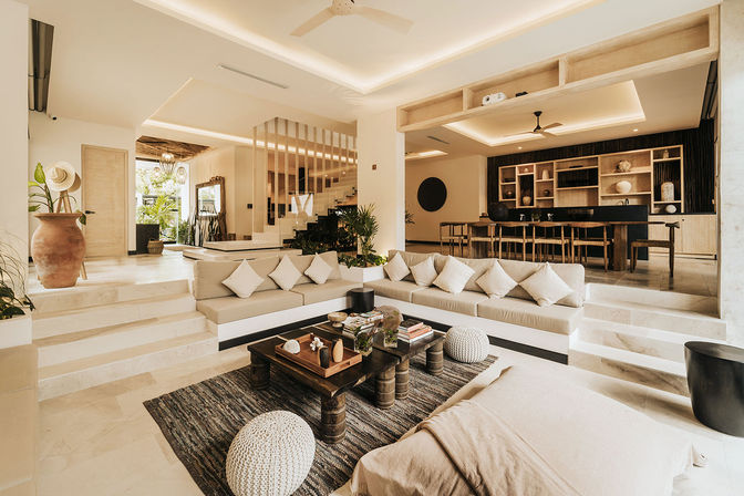 Sunlit resort-style open-plan living room with sunken built-in neutral sofa and throw pillows, wooden coffee table on textured rug, round knit poufs, terracotta vase and plants, steps up to a long wooden dining table and open shelving—modern tropical villa interior.