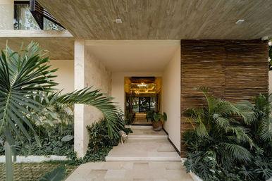 Inviting modern tropical villa entrance with stone steps and covered entryway, lush palm landscaping, a wooden slat wall and large potted plant leading to a glass door with warm interior lighting.