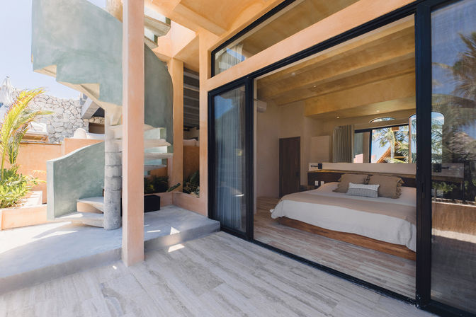 Sunlit modern coastal villa bedroom with sliding glass doors to a terrace, concrete spiral staircase, tropical plants and palm-tree views