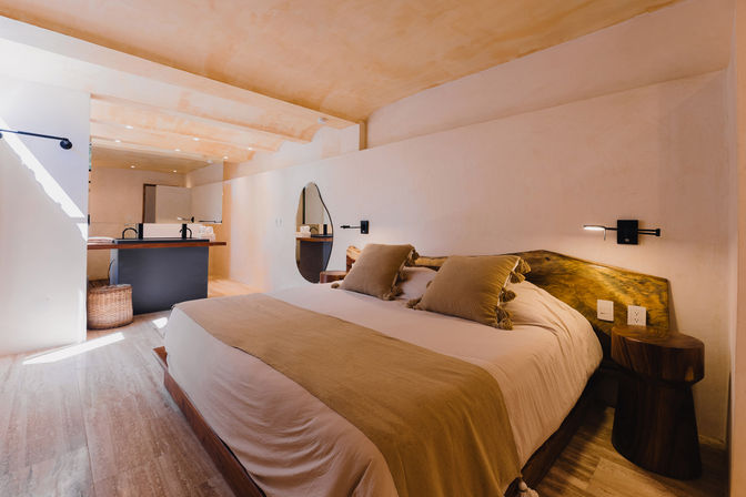 Sunlit modern rustic bedroom with king bed, neutral linens and tassel pillows, live-edge wood headboard and round wooden side table, wall-mounted reading lights, wood floors and open-plan vanity with round mirror — cozy resort-style suite.