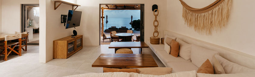 Beachfront open-plan living room with built-in beige sofa, wooden tables, woven wall hanging and wall-mounted TV, opening onto a patio with round dining table and ocean view