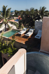 Tropical rooftop deck with a small plunge pool and three white cushioned loungers on a wooden terrace, framed by palm trees and a distant ocean view.
