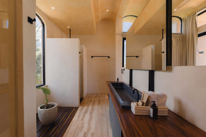 Sunlit minimalist bathroom with warm plaster walls, arched windows, travertine floor, wood countertop and long black trough sink with dual matte-black faucets, potted plant and private shower stalls