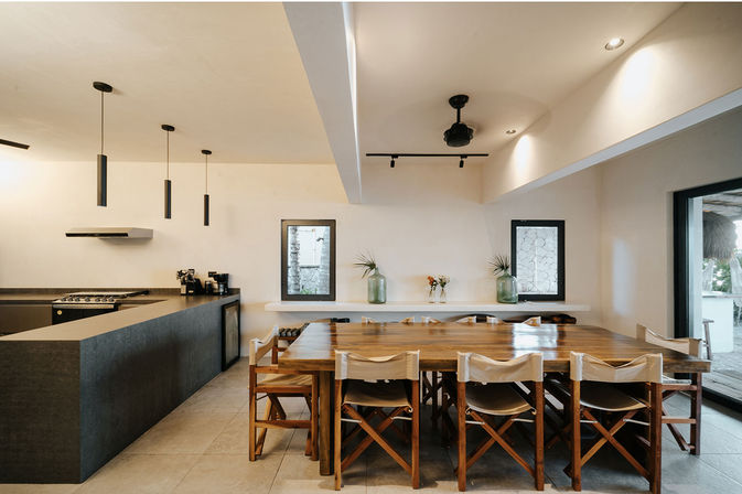 Sunlit open-plan modern kitchen and dining area in a coastal villa — long wooden table with canvas sling chairs, dark stone kitchen island with pendant lights, minimalist white walls, small windows and sliding patio door