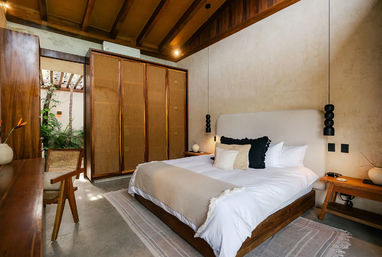 Sunlit tropical-inspired bedroom with exposed wooden beams, woven-panel wardrobe, low wooden bed dressed in neutral linens and textured pillows, black pendant bedside lights, wooden desk and chair, and a small indoor courtyard with green plants visible through an open doorway.