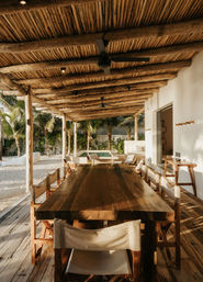 Sunlit beachfront patio with a long rustic wooden dining table and canvas chairs under a reed-and-log pergola with ceiling fans, surrounded by palm trees, sand and a small plunge pool.