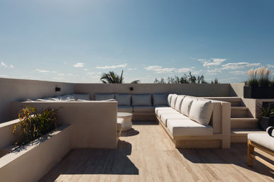 Sun-drenched modern rooftop terrace with built-in L-shaped cream sectional, plush white cushions, potted plants and a clear blue sky — stylish outdoor lounge seating