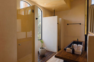 Modern tropical bathroom with warm plaster walls, glass walk-in shower and arched window showing palm fronds, wooden slatted floor mat and potted plant, wood countertop with black sink, rolled towels and toiletries in natural light.