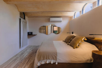 Cozy minimalist bedroom with a large bed in neutral linens and tassel-trim throw, wooden stump nightstand, arched clerestory windows, wavy decorative mirror and wall-mounted air conditioner