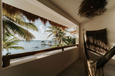 Sunlit tropical balcony with a hammock, palm-fringed ocean view and two thatched overwater huts on a wooden pier